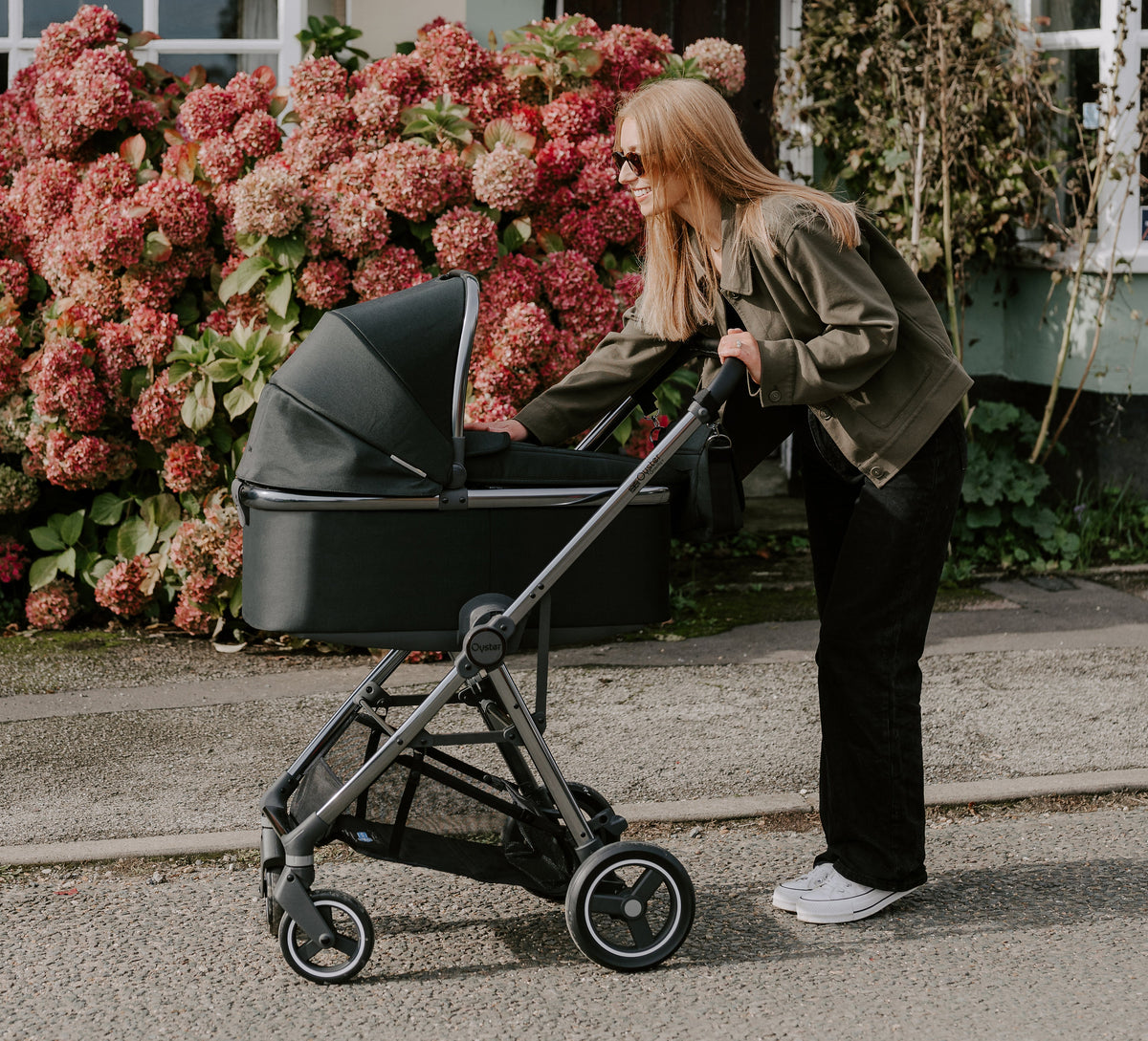 Woman pushing a stroller in front of a house with pink flowers.