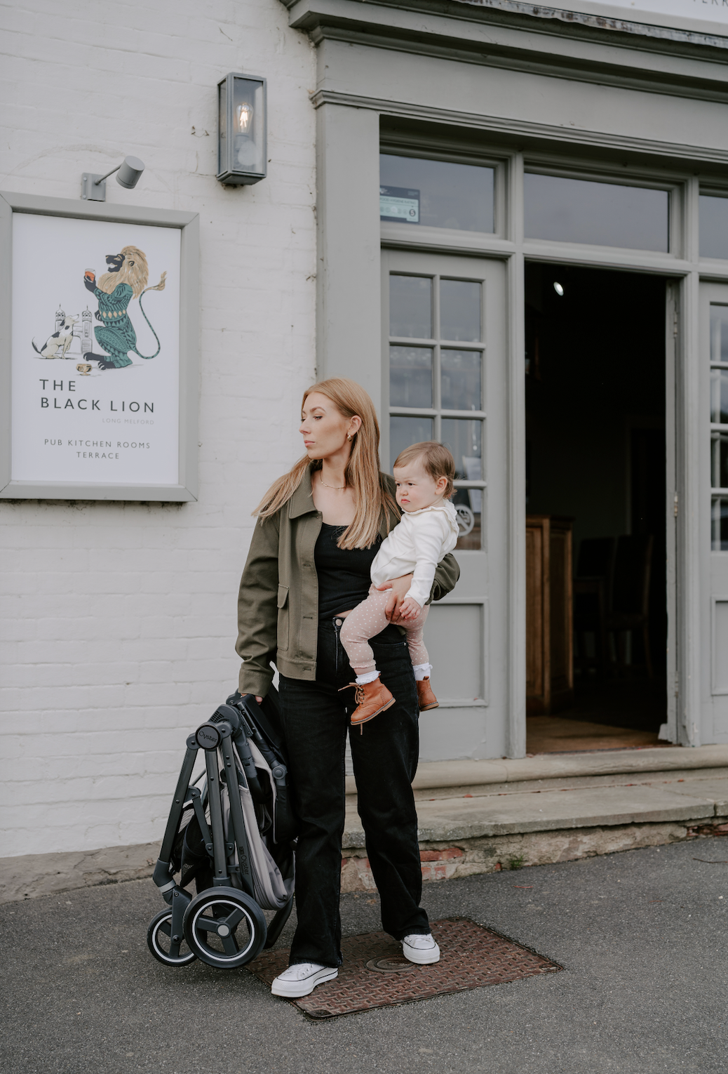 Woman holding a child and pushing a stroller outside a building with a sign for 'The Black Lion'.
