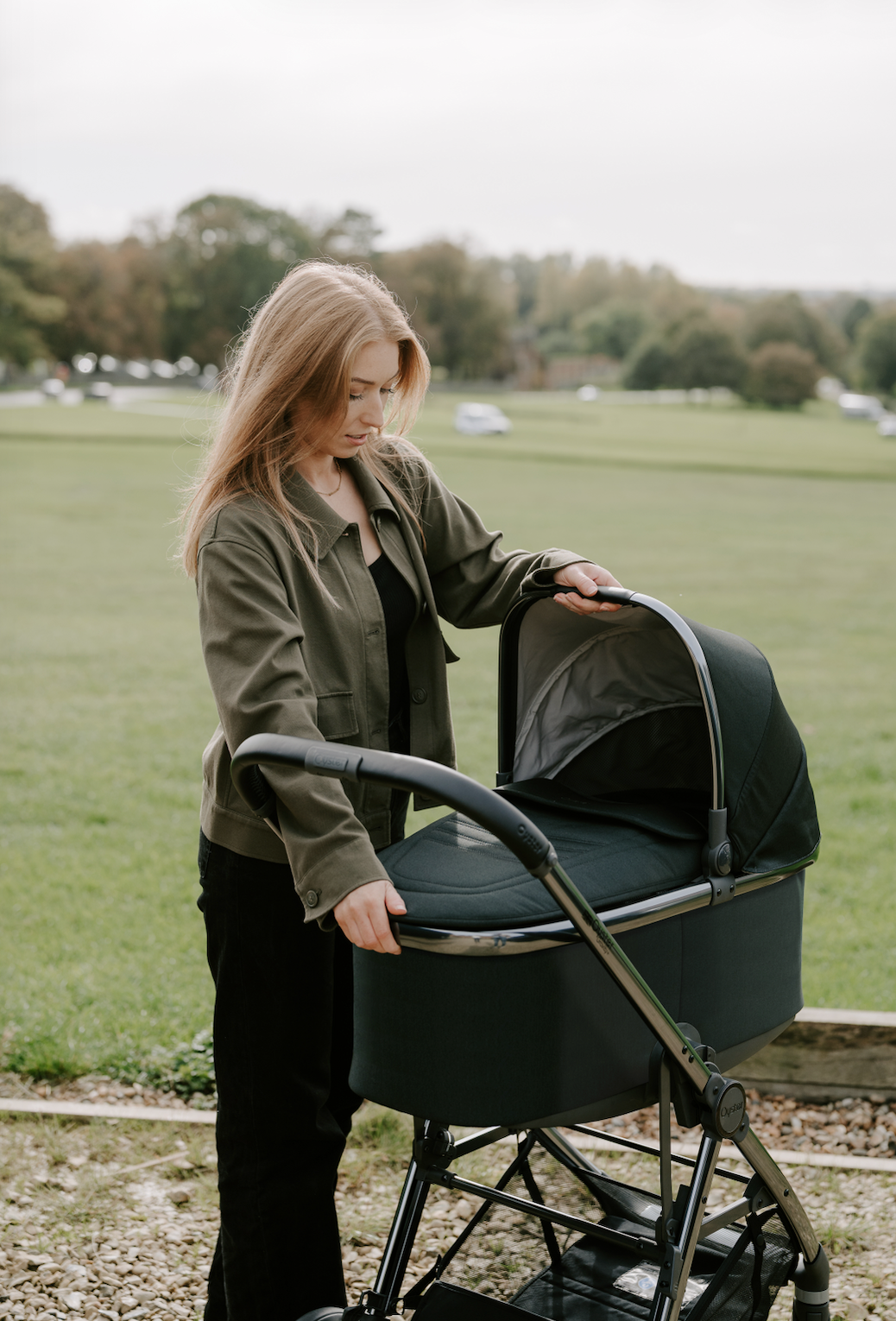 Woman pushing a green pram in an open field with trees in the background
